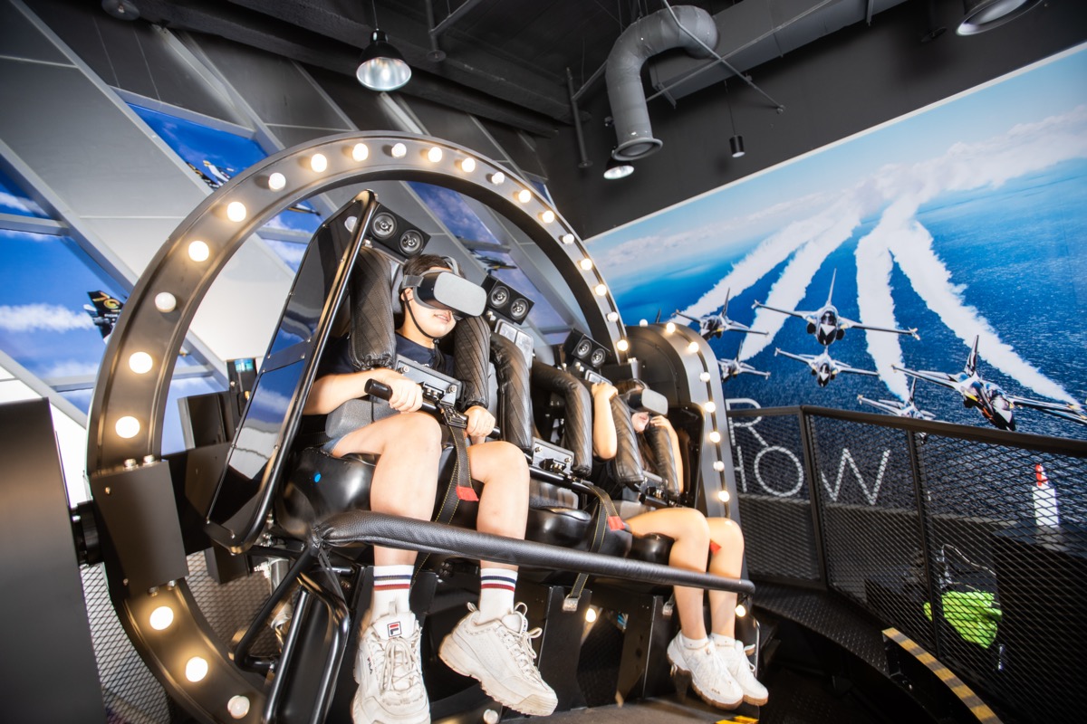 VR 헤드셋을 쓴 체험객들이 블랙이글스탑승체험 시뮬레이터 좌석에 앉아 있고, 뒤편 벽면에는 블랙이글스 편대비행 사진이 보인다
